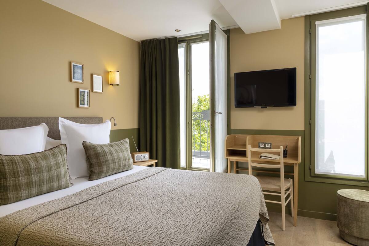 Ein Hotelzimmer mit einem großen Bett, einem Flachbildfernseher, einem Schreibtisch mit Stuhl und einem fenster mit Vorhängen.