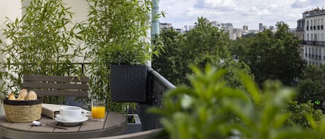 A balcony with a wicker chair, a wicker basket with bread, a white cup, and a glass of orange juice.