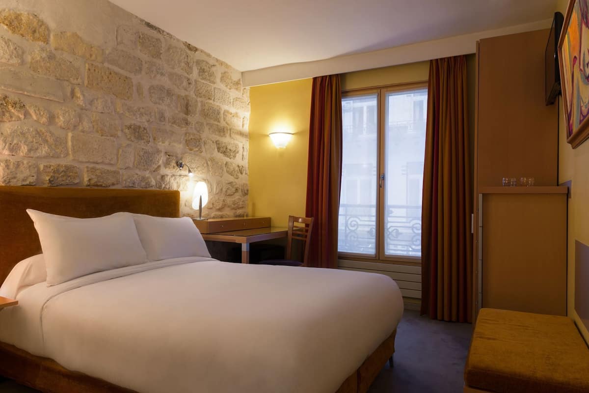 A hotel room with a stone wall, a bed with white linens, a desk with a lamp, a window with curtains, and a door with a mirror.