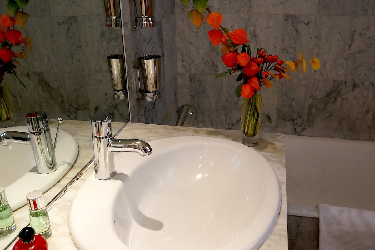 A bathroom with a white sink, two chrome faucets, a mirror, and a vase with artificial flowers.