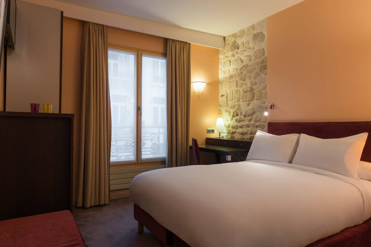 A hotel room with a bed, a desk, a chair, a lamp, and a stone wall.