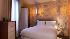 A neatly made bed in a room with stone walls, a wooden dresser, and a small table with a lamp. - Hôtel Danemark (Paris)