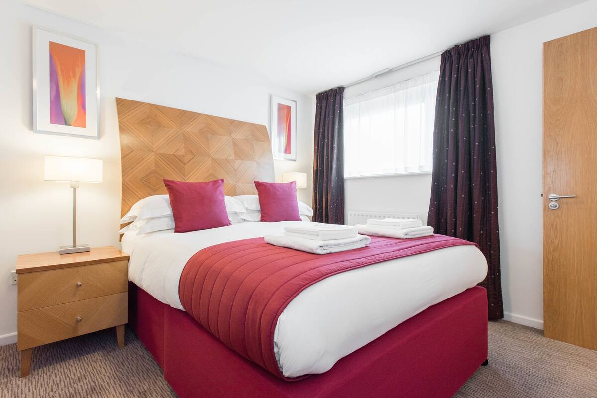 Een hotelkamer met een bed, nachtkastje, lamp, raam met gordijnen en een deur.