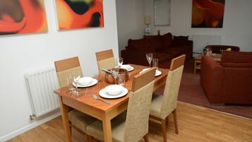 A dining area with a wooden table set for four, surrounded by wicker chairs. A brown sofa is in the background, and abstract art is on the walls.