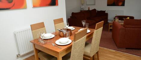 Un comedor con una mesa de madera dispuesta para cuatro personas, rodeada de sillas de mimbre. En el fondo, un sofá marrón y obras de arte abstracto en las paredes.