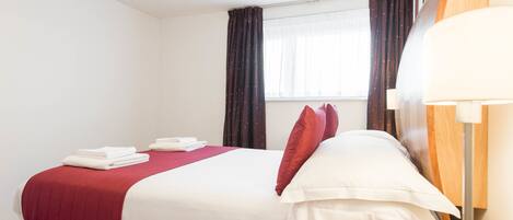 A neatly made bed with white and red bedding, a wooden floor, a window with curtains, and a bedside lamp.