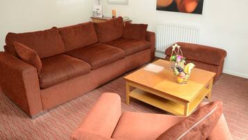 A living room with a brown sofa, a wooden coffee table with a vase of fruit, and a radiator on the wall.