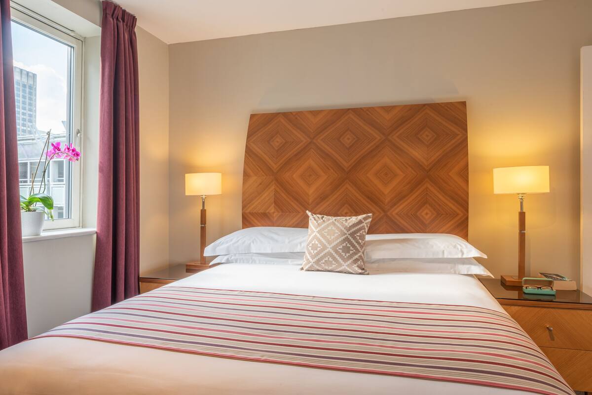 A hotel room with a bed, two bedside lamps, a window with curtains, and a potted plant.