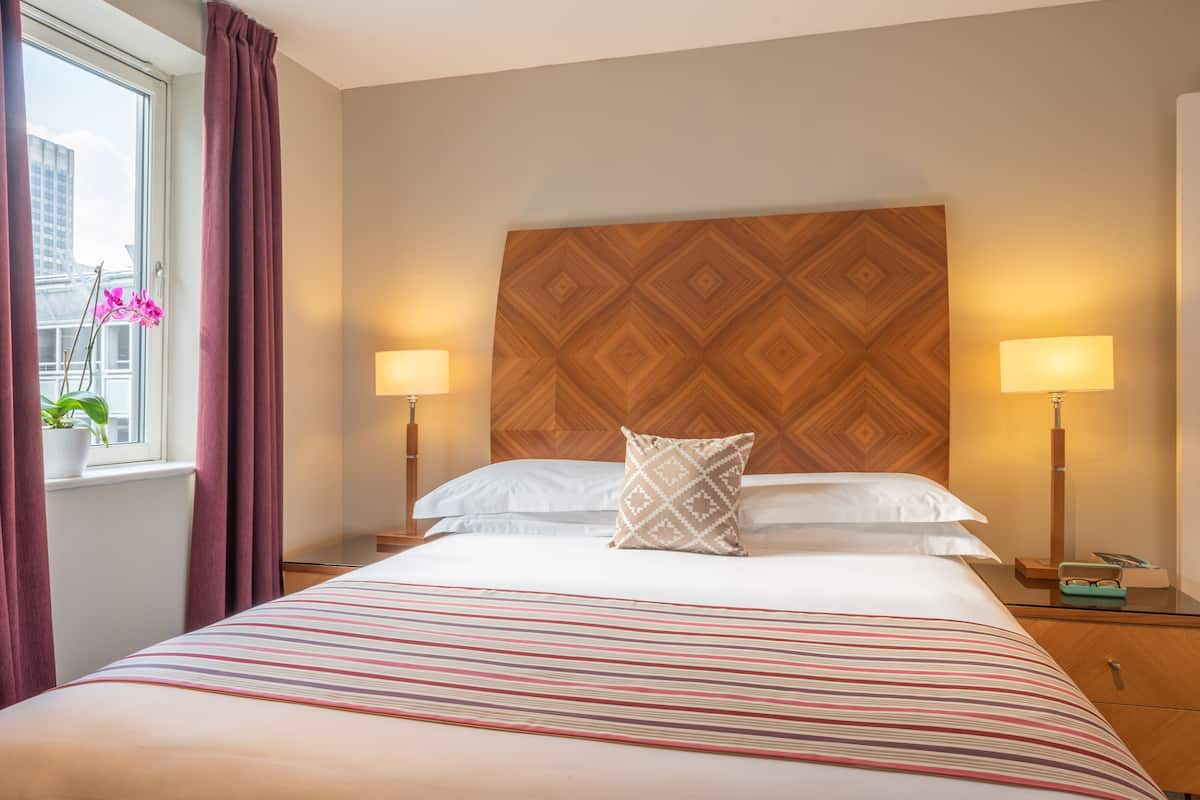 A hotel room with a bed, two bedside lamps, a window with curtains, and a potted plant.