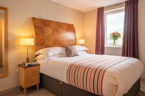 Una habitación de hotel con una cama, dos lámparas de noche, una mesita de noche, una ventana con cortinas y vistas a la ciudad.