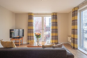 Ett vardagsrum med en soffa, en tv på ett tv-bord, en vas med blommor och gardiner.