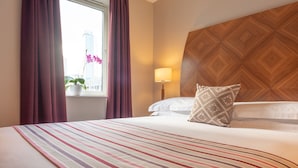 Hotellihuone, jossa on suuri sänky, ikkuna verhoilla, ruukkukasvi ja puinen pääty.