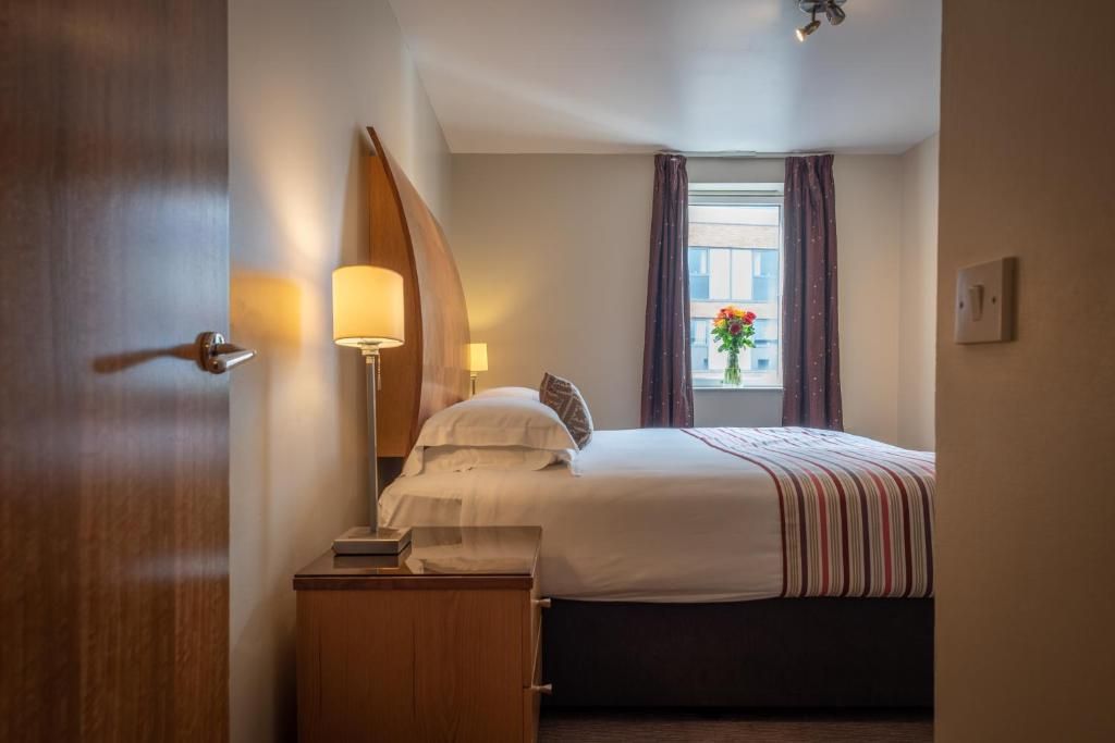 Una camera d'albergo con un letto, comodino, lampada e finestra con tende.