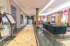 Um hall de hotel com piso de mármore, balcão de recepção, área de estar e grandes janelas.