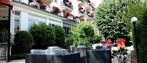 An outdoor seating area with wicker furniture, a large tree, and a building with balconies.