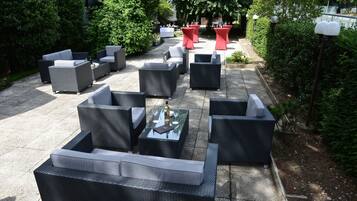 An outdoor seating area with wicker furniture and red tables, surrounded by greenery.