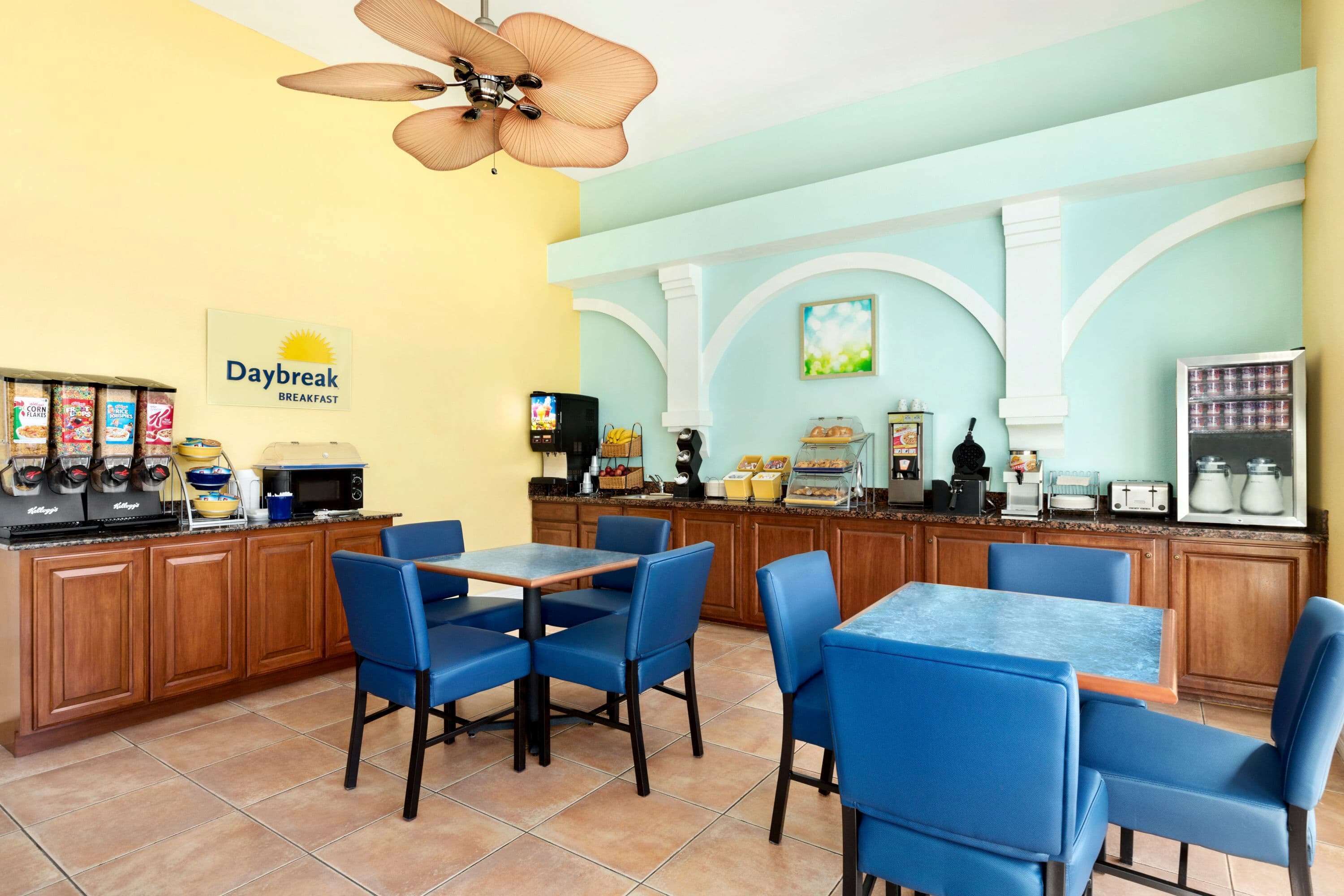 A breakfast area with a sign reading "Daybreak Breakfast," featuring a kitchen counter with coffee machines, a microwave, and a toaster. There are blue chairs and tables arranged for dining.