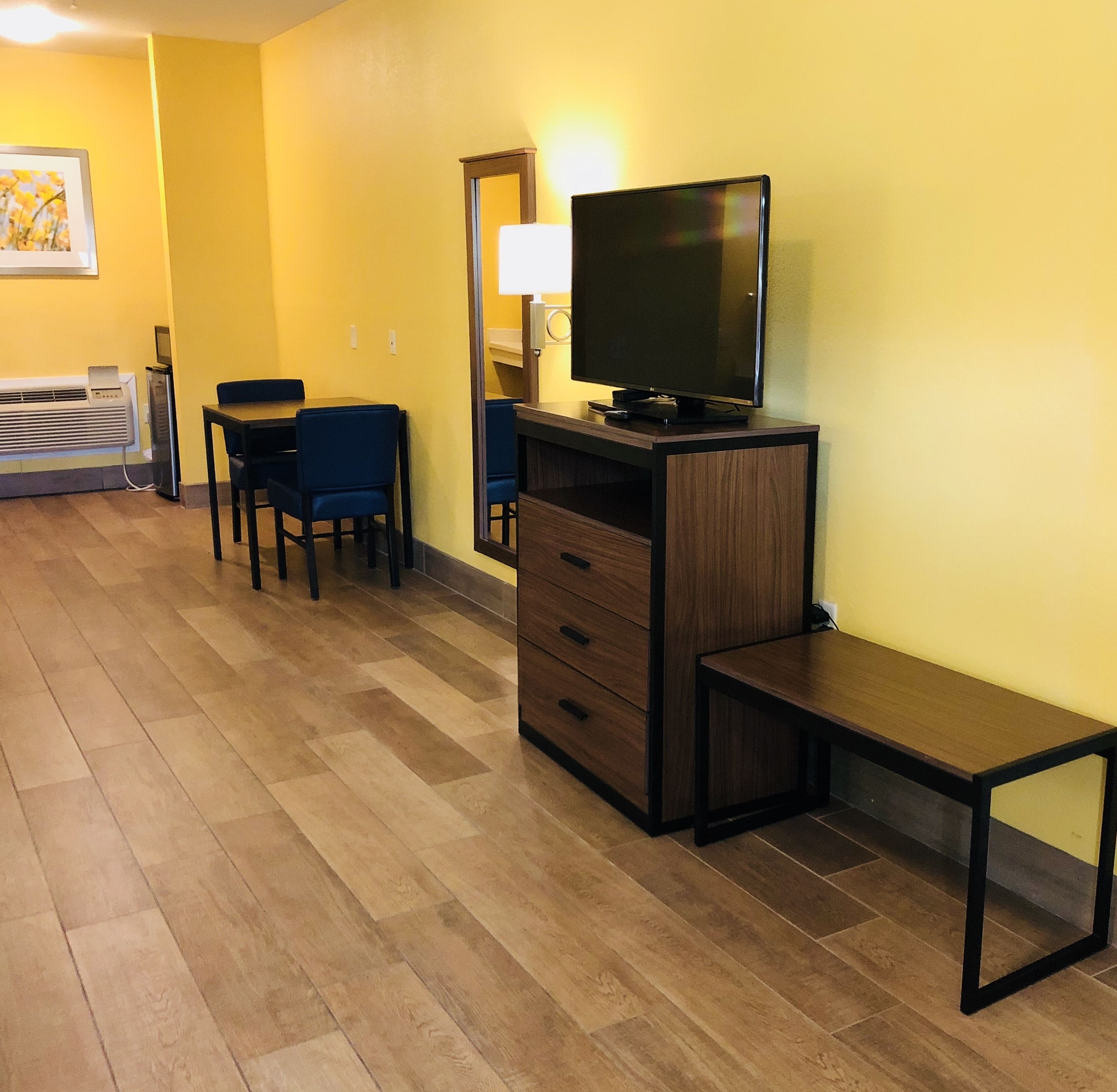A hotel room with a wooden floor, a TV on a wooden stand, a desk with a chair, and a bench.