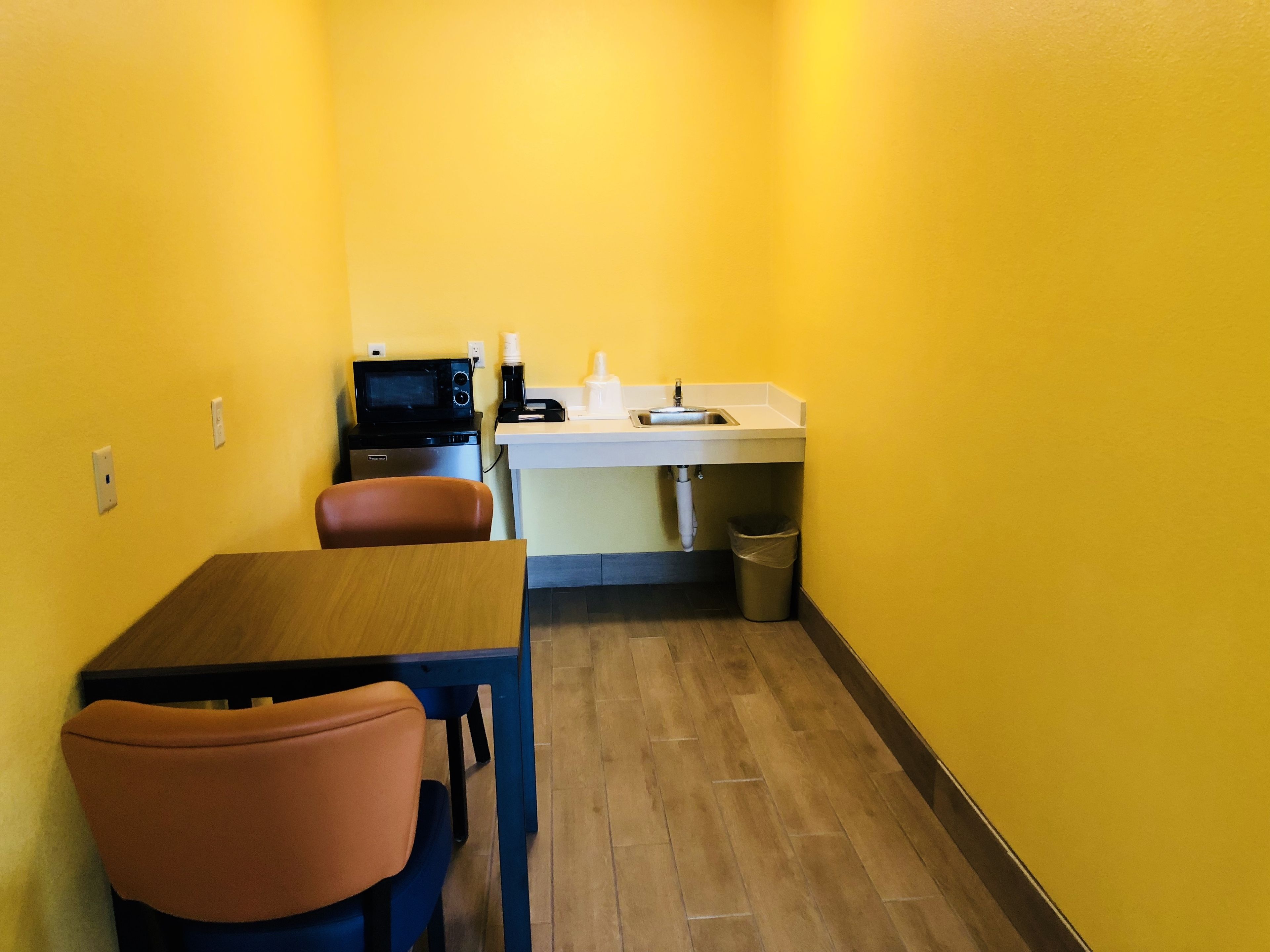 A small kitchenette with a microwave, coffee maker, and sink. There is a wooden table and two chairs.