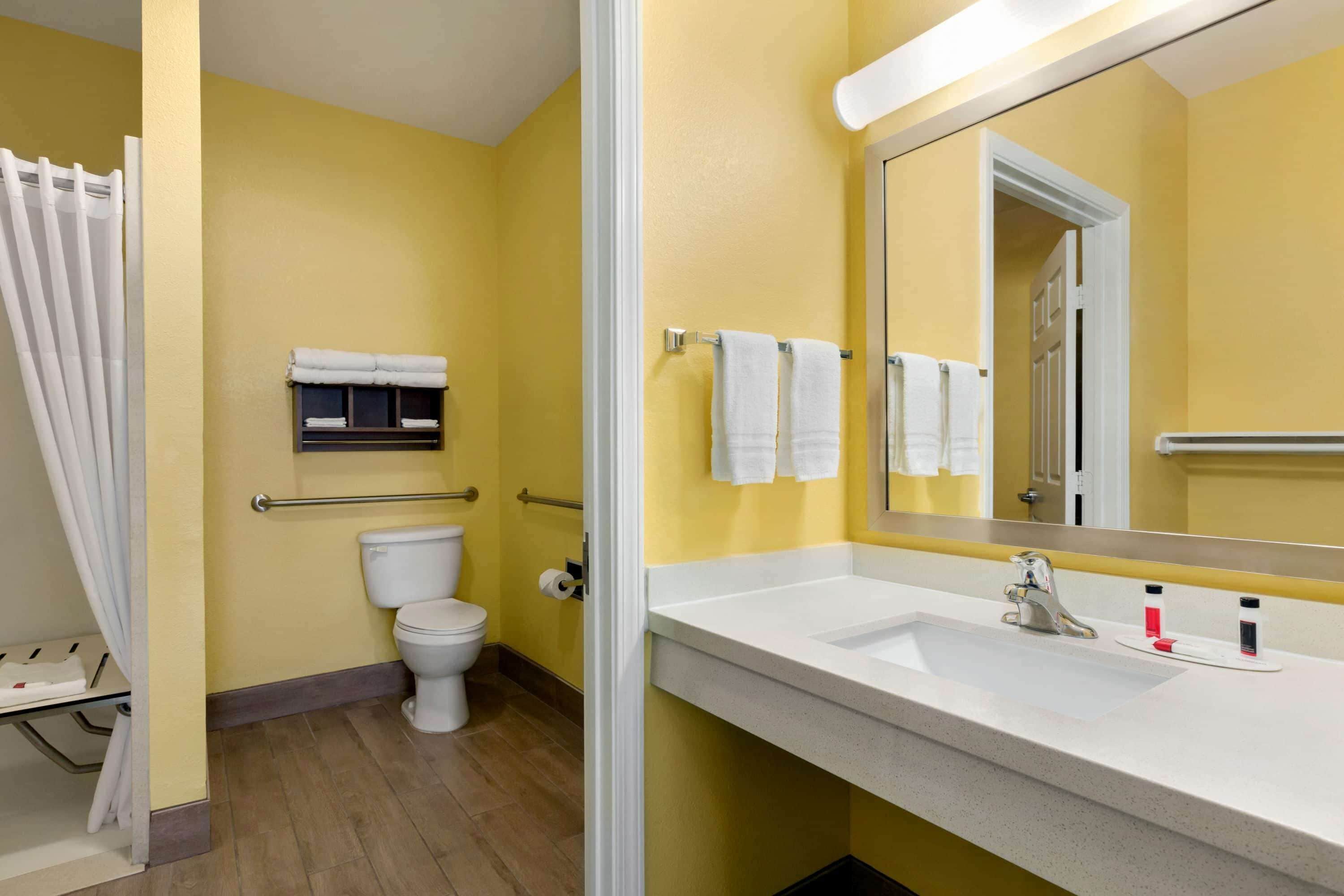 Un baño con pared amarilla, inodoro blanco y un espejo grande sobre un lavabo con dos dispensadores de jabón.