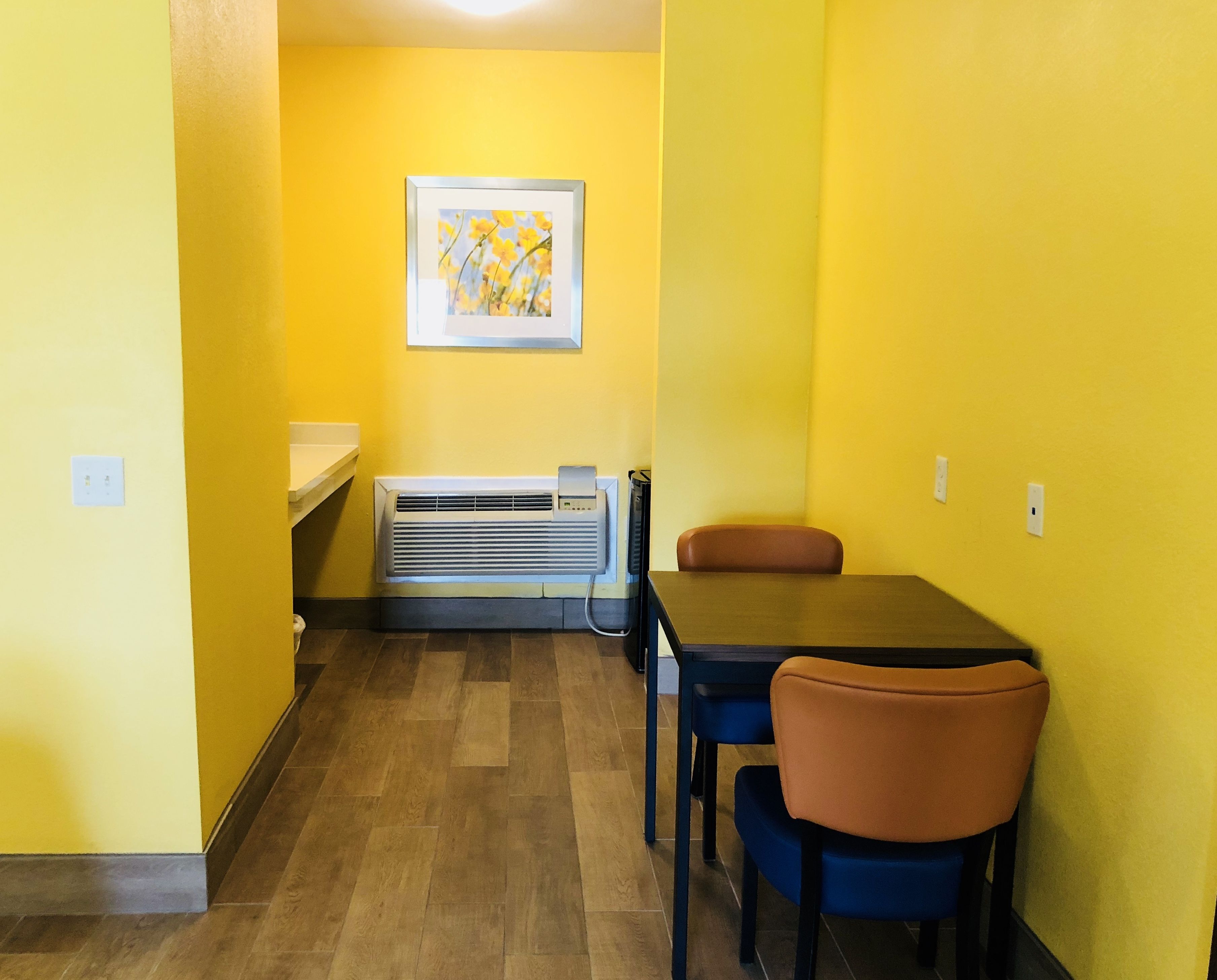 Une petite pièce, bien éclairée, aux murs jaunes, avec un plancher en bois, une table et deux chaises, un radiateur et un tableau encadré accroché au mur.