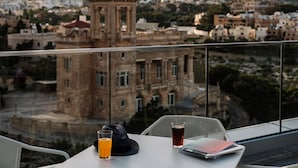 Un balcón con una mesa, un vaso de zumo de naranja, un vaso de líquido oscuro y un libro.