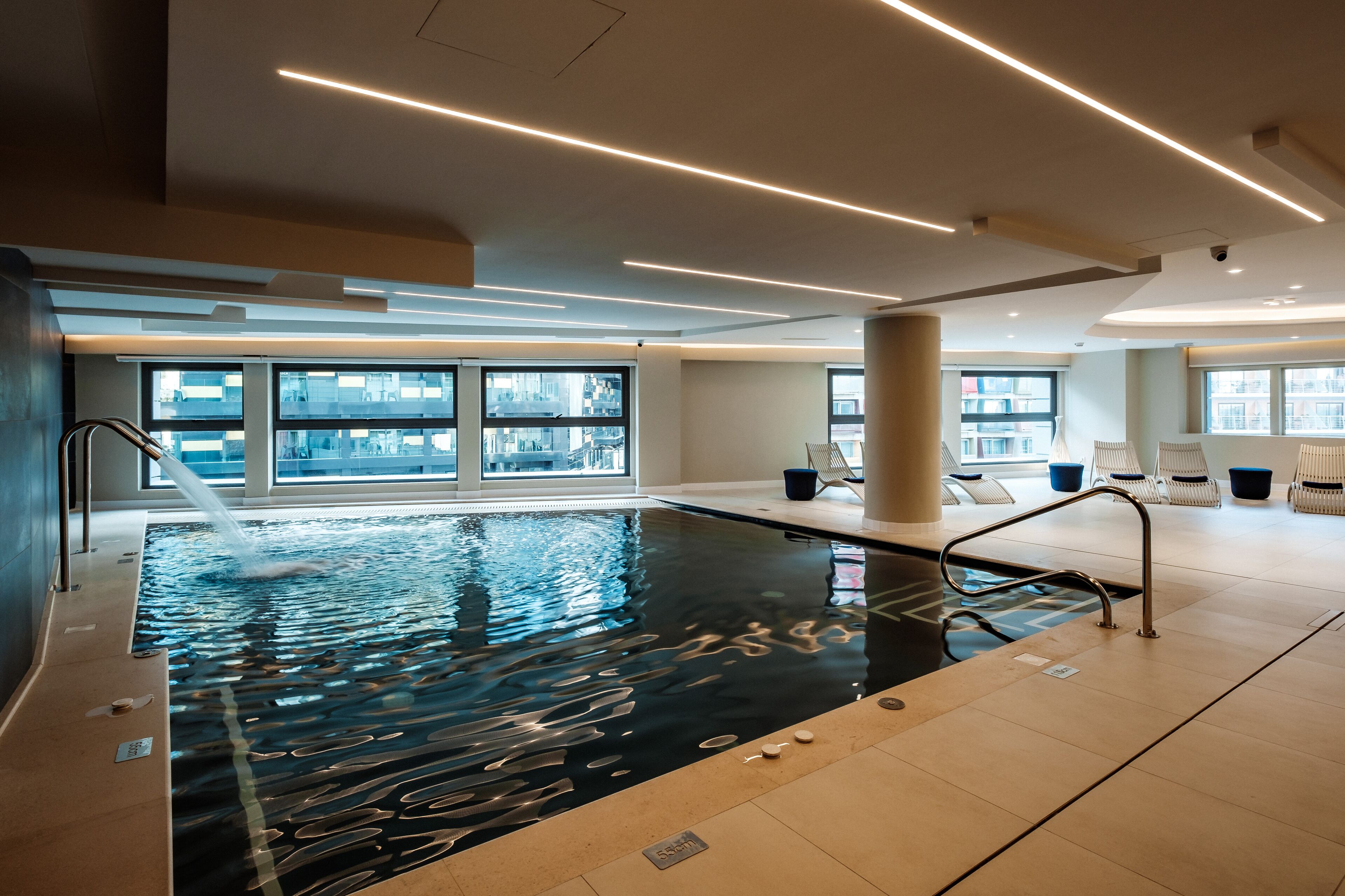 An indoor swimming pool with clear windows, modern lighting, and lounge chairs.