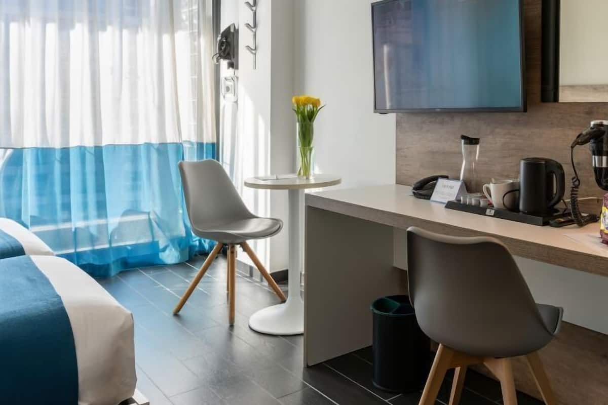 A hotel room with a bed, a desk with a computer, a chair, a small table with a vase, and a TV on the wall.