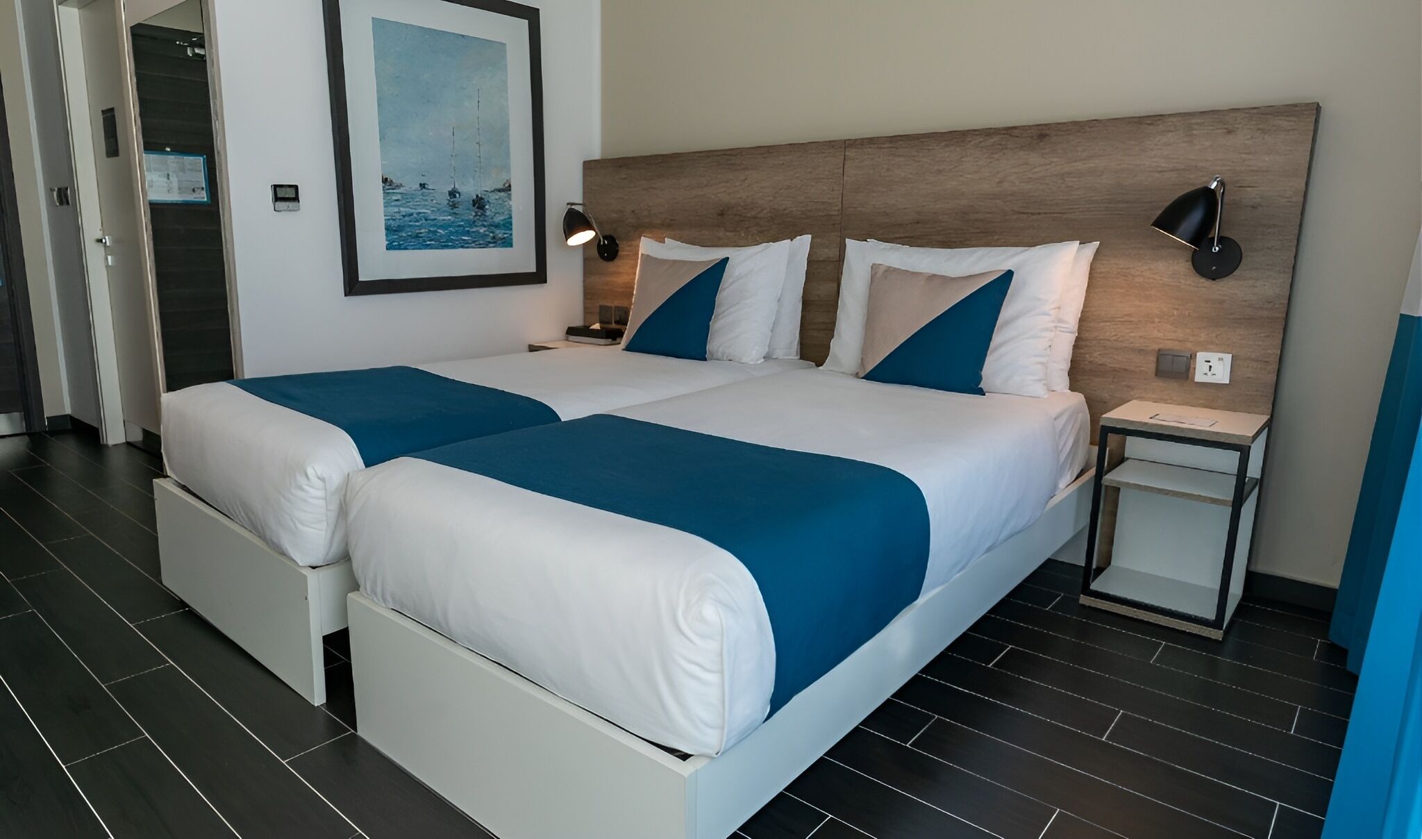 A double bed with white and blue bedding, a wooden headboard, two bedside tables with lamps, and a framed picture on the wall.