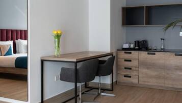 A modern hotel room with a bed, a desk with two chairs, a small kitchen area, and a large mirror.