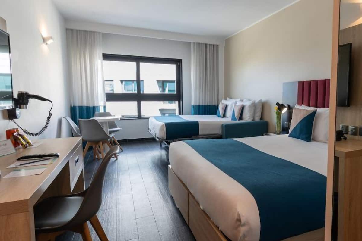 A hotel room with a desk, two beds, a large window, and a TV.