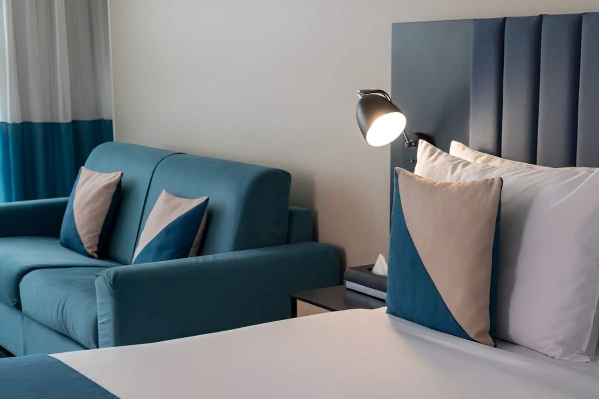 A hotel room with a bed, a blue sofa, a bedside table, and a lamp.