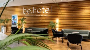 A modern hotel reception area with a wooden wall, a green sofa, black chairs, and potted plants. - be.HOTEL (St. Julian's)