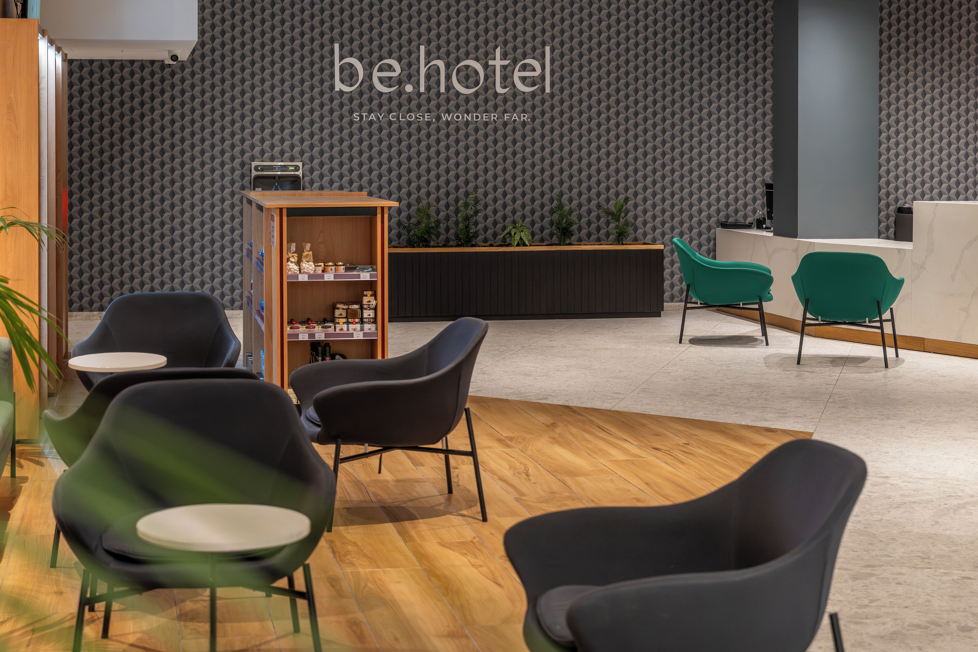 Un espace d’accueil d’hôtel au design moderne, comprenant un comptoir d’accueil, des chaises vertes, un meuble en bois contenant des articles de toilette et un mur orné d’un motif géométrique.