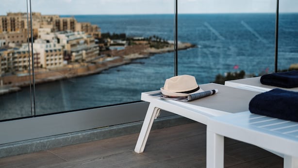 A balcony with a view of the sea and buildings, a white table with a hat and a towel, and a wooden floor. - be.HOTEL (St. Julian's)