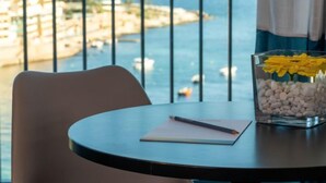 A round table with a notebook and pen, a glass vase with yellow flowers, and a view of the sea and buildings through a window with metal bars. - be.HOTEL (St. Julian's)