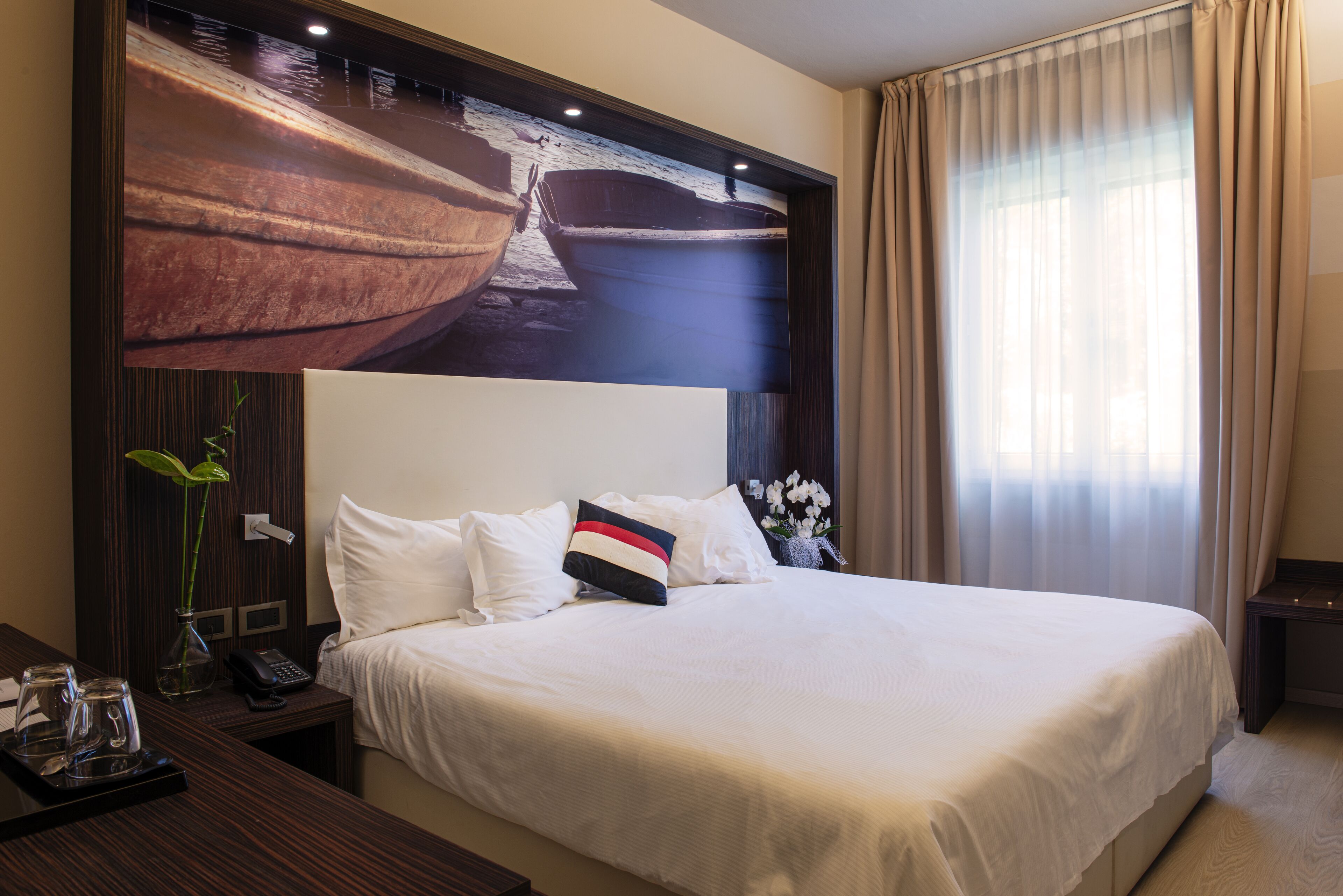 Habitación de hotel con una cama grande, una mesita de noche con un teléfono y un jarrón de flores, y una obra de arte en la pared que representa barcos.