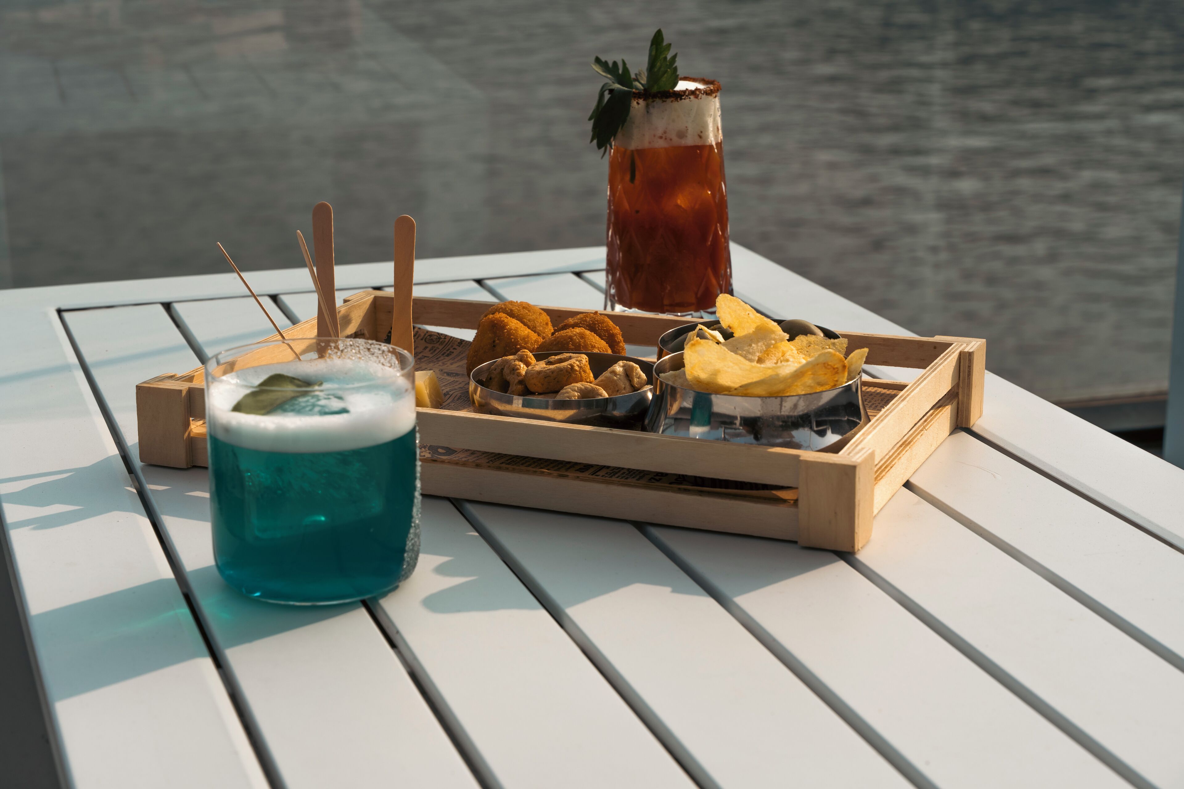 Una mesa con una bandeja de comida y bebidas, que incluye una bebida azul, alimentos fritos y un vaso de té helado, frente a una masa de agua.