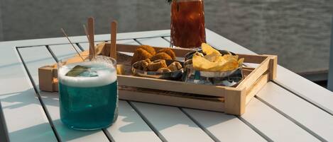 Una mesa con una bandeja de comida y bebidas, que incluye una bebida azul, alimentos fritos y un vaso de té helado, frente a una masa de agua.