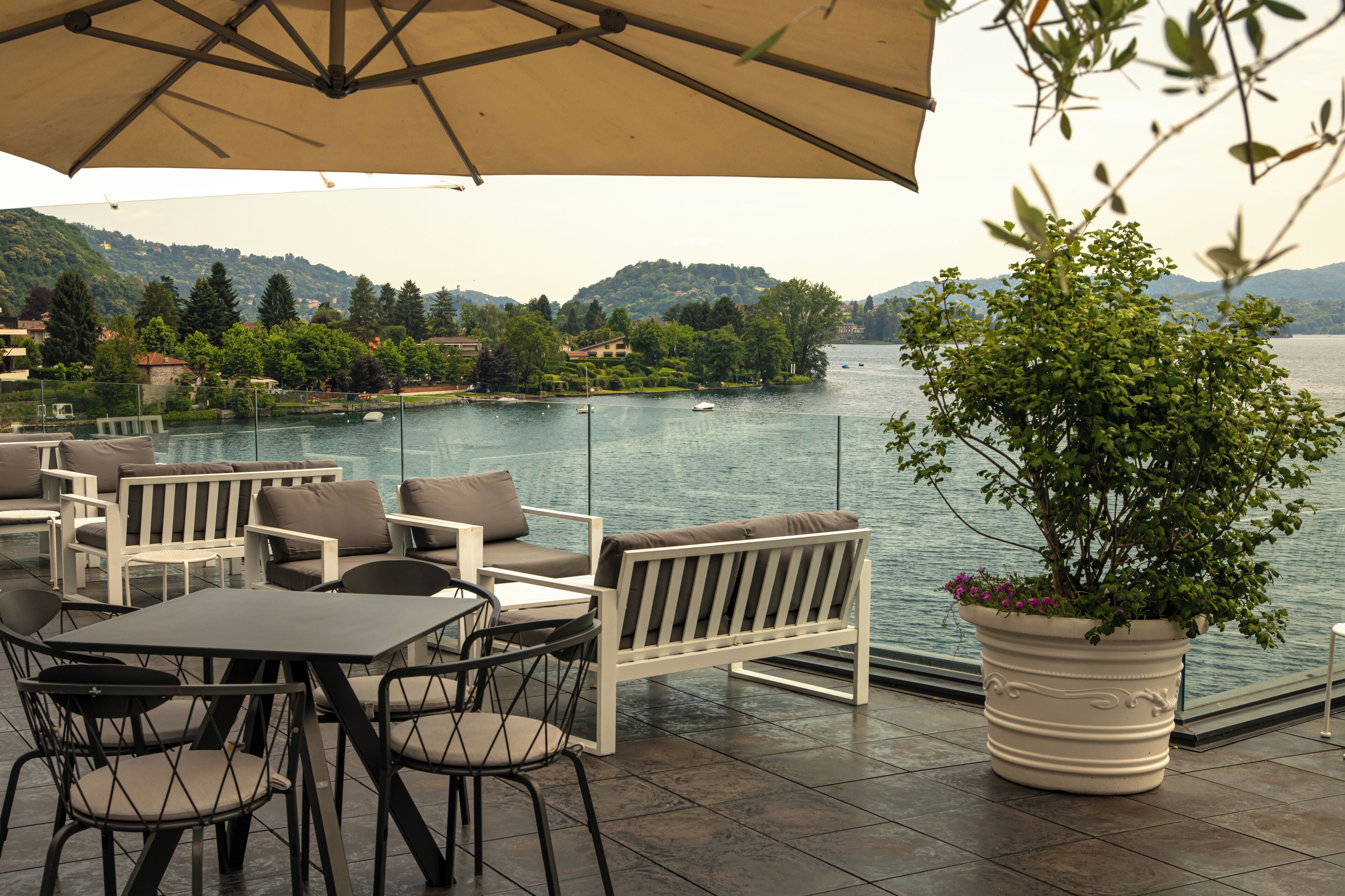 Un espacio al aire libre con mesas y sillas, una planta en maceta, y vistas a un lago y colinas.