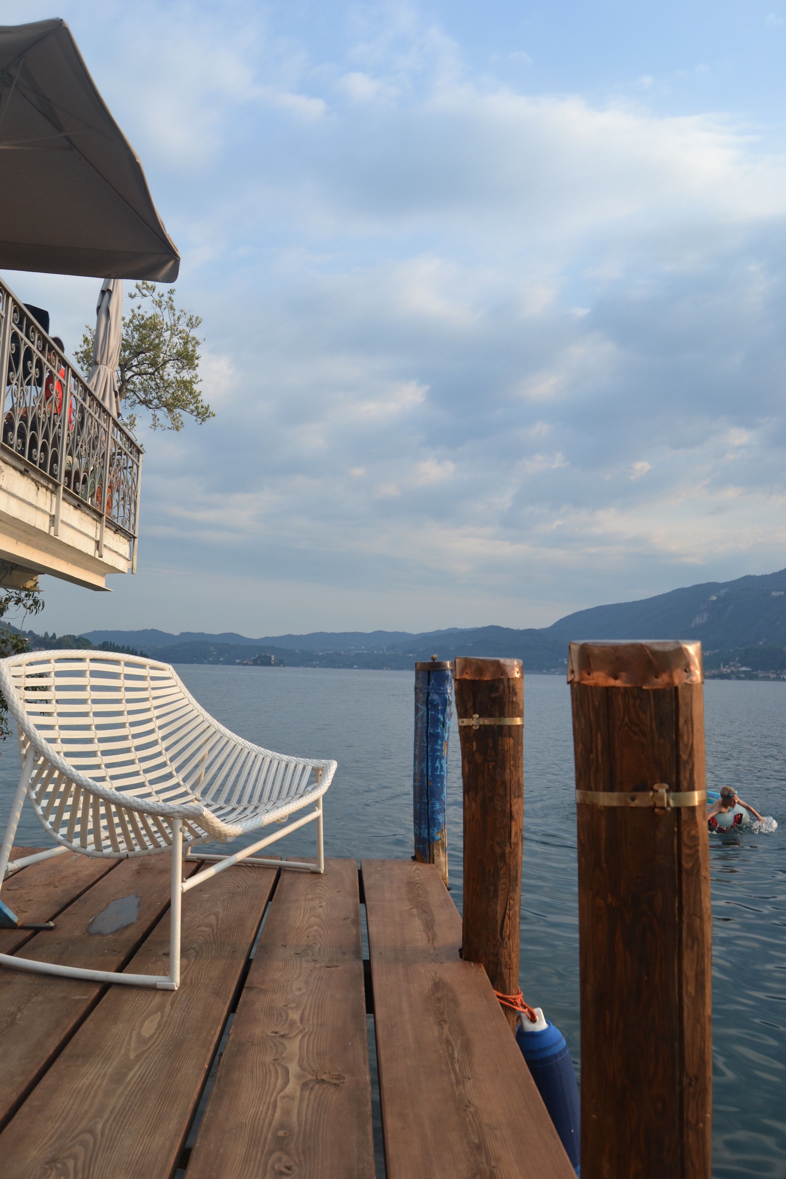 Une terrasse en bois avec une chaise et une table en métal peintes en blanc, donnant sur un lac paisible, avec des montagnes en arrière-plan.