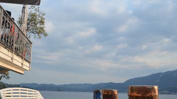 A wooden deck with a white metal chair and a table, overlooking a calm lake with mountains in the background.
