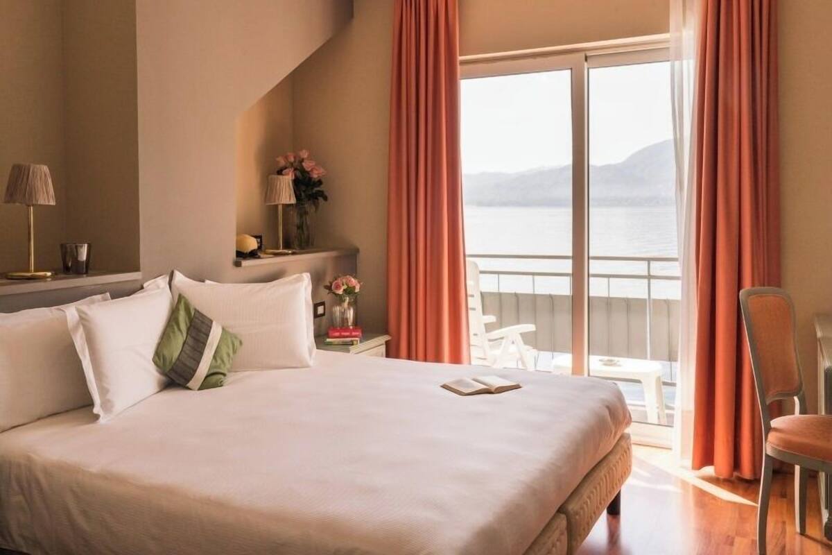 Une chambre d’hôtel avec un grand lit, une table de chevet avec une lampe, un vase de fleurs et une vue sur la mer depuis un balcon.