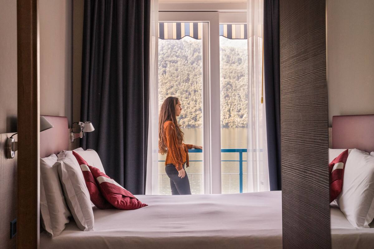 Une chambre d’hôtel avec un lit, une table de chevet, une lampe et une vue sur un balcon doté d’une balustrade.