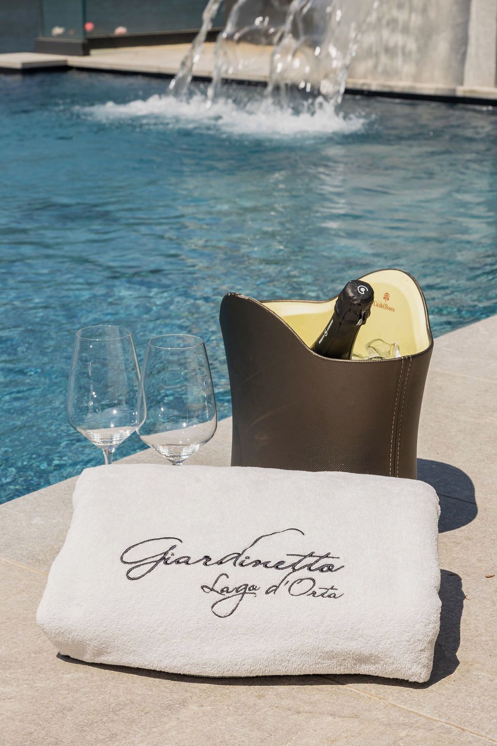 Una botella de champán en un cubo marrón, dos copas de vino vacías y una toalla con la inscripción "Giardinetto Lago d'Ota", junto a una piscina con una fuente de agua al fondo.