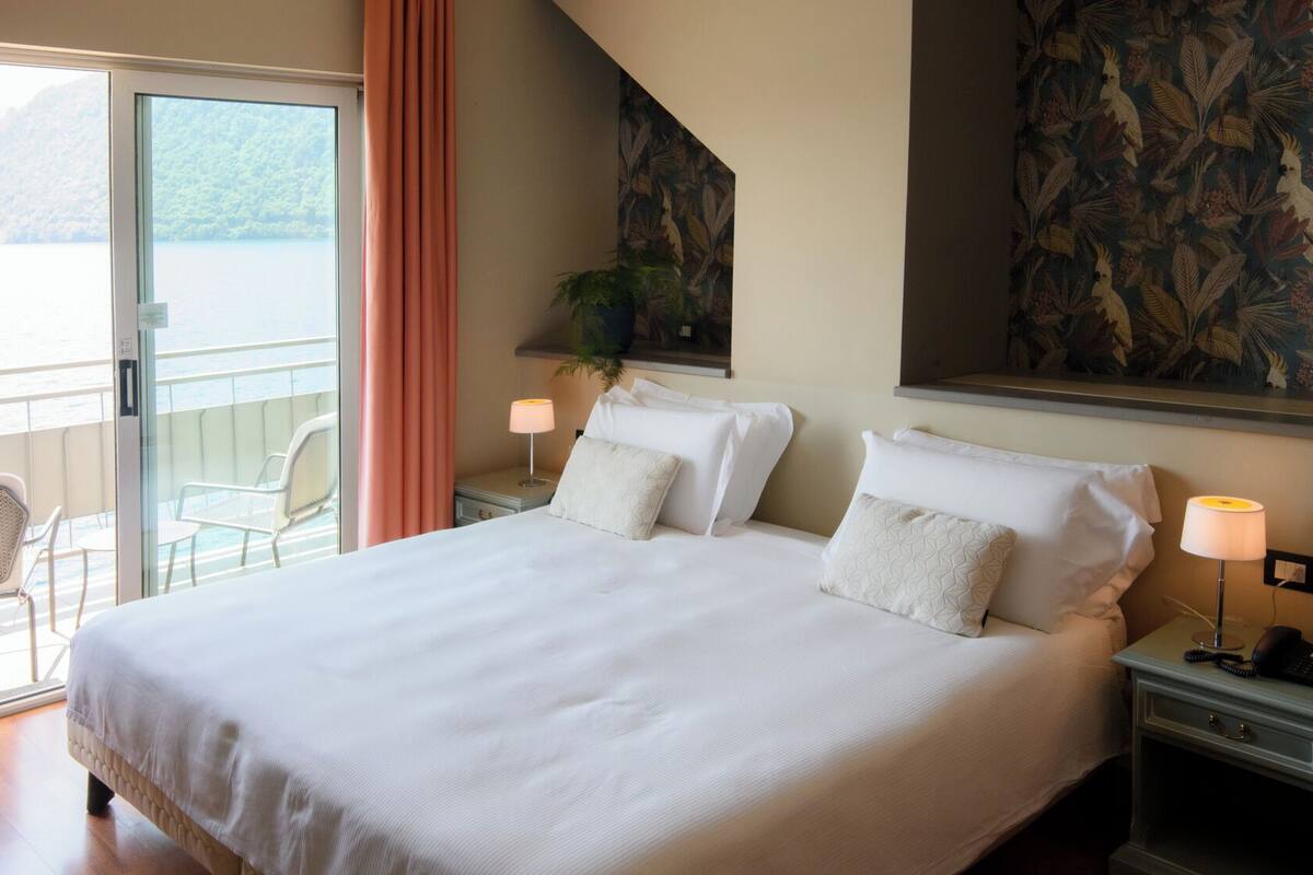 Une chambre d’hôtel avec un grand lit, deux tables de chevet avec des lampes, une vue sur un balcon avec des chaises et une fresque murale sur le thème des tropiques.