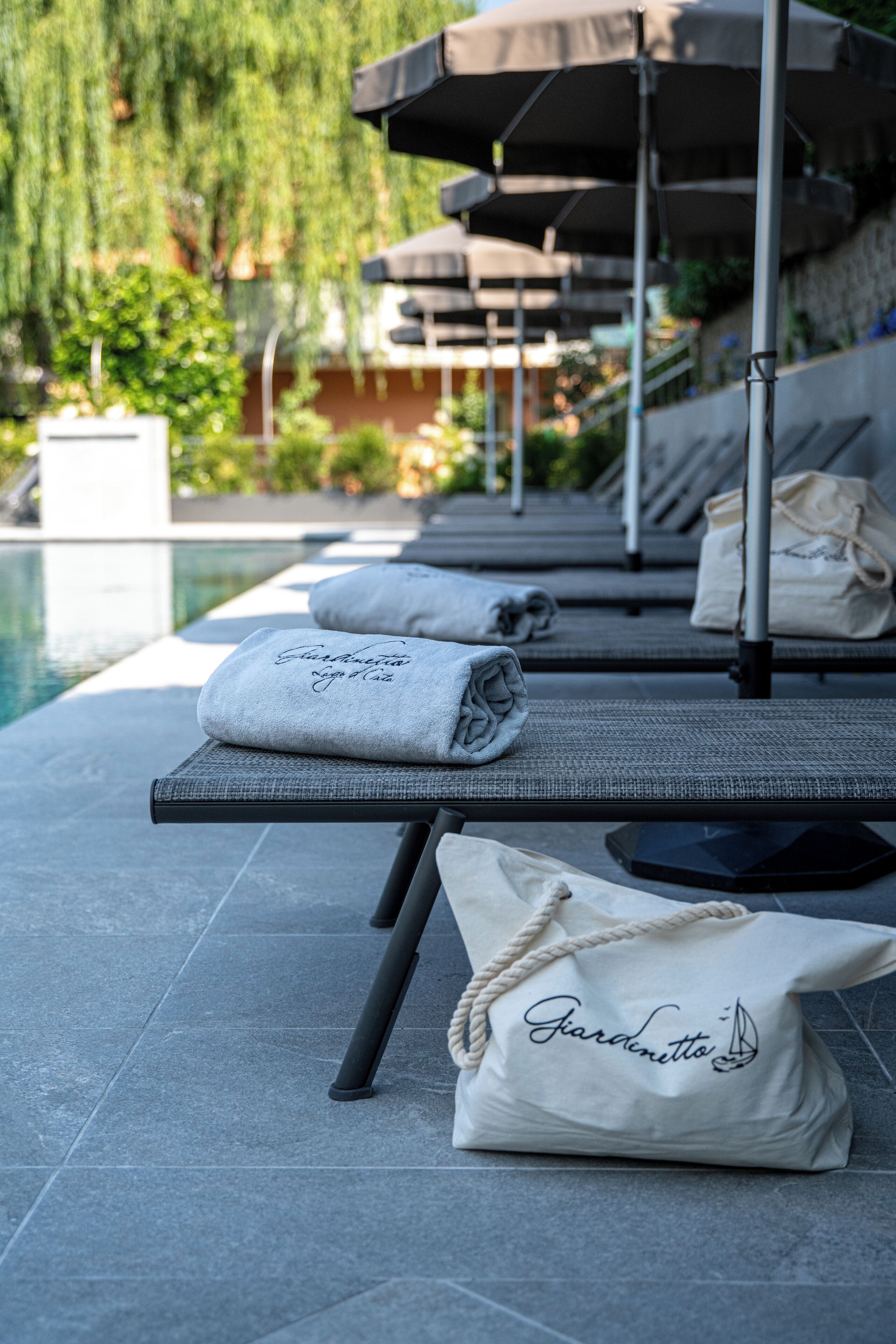 A poolside lounge area with sun loungers, umbrellas, and towels with 'Giardinetto' branding.