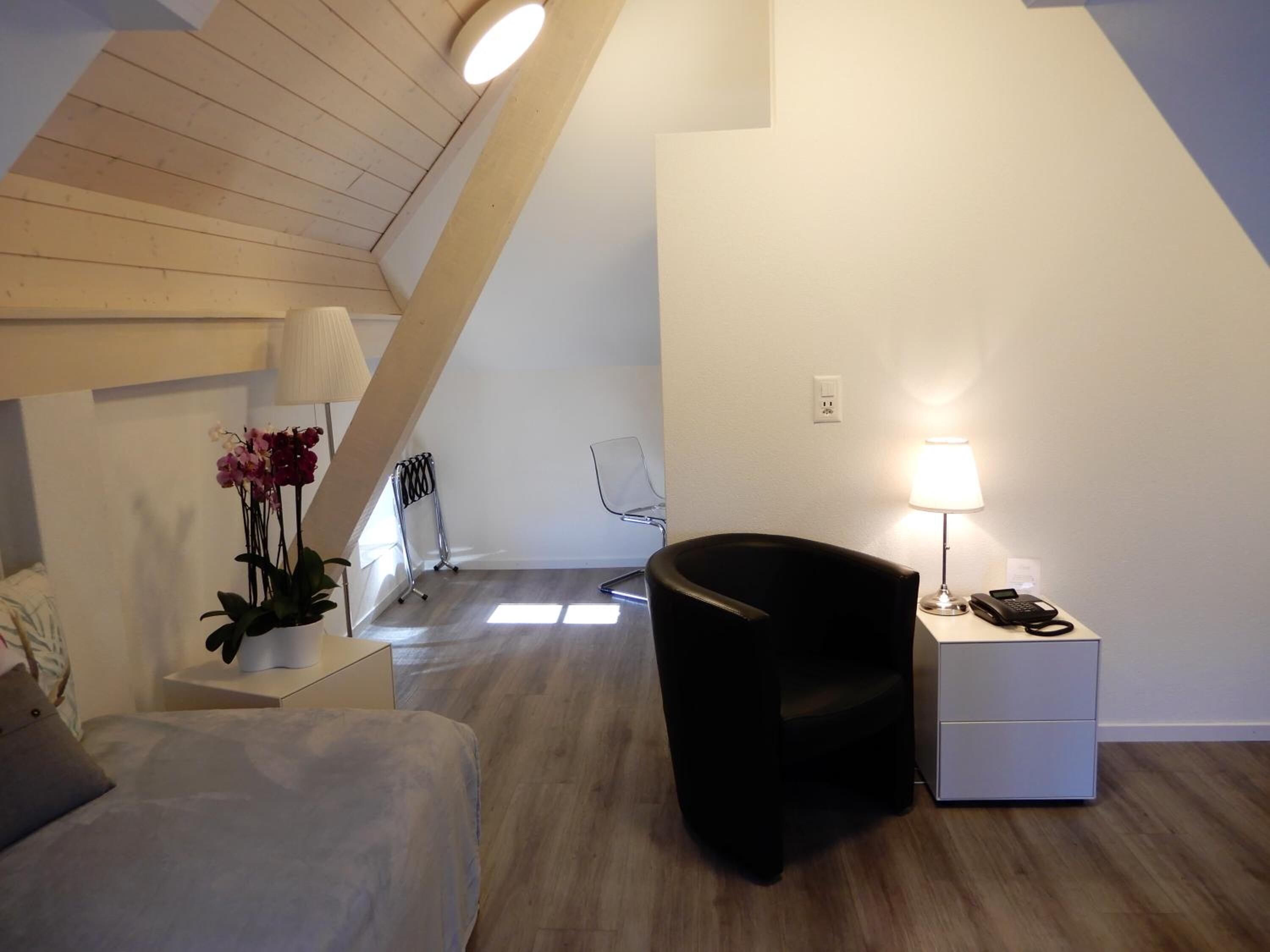 A modern bedroom with a sloped ceiling, a black chair, a bedside table with a lamp, and a small desk with a phone.