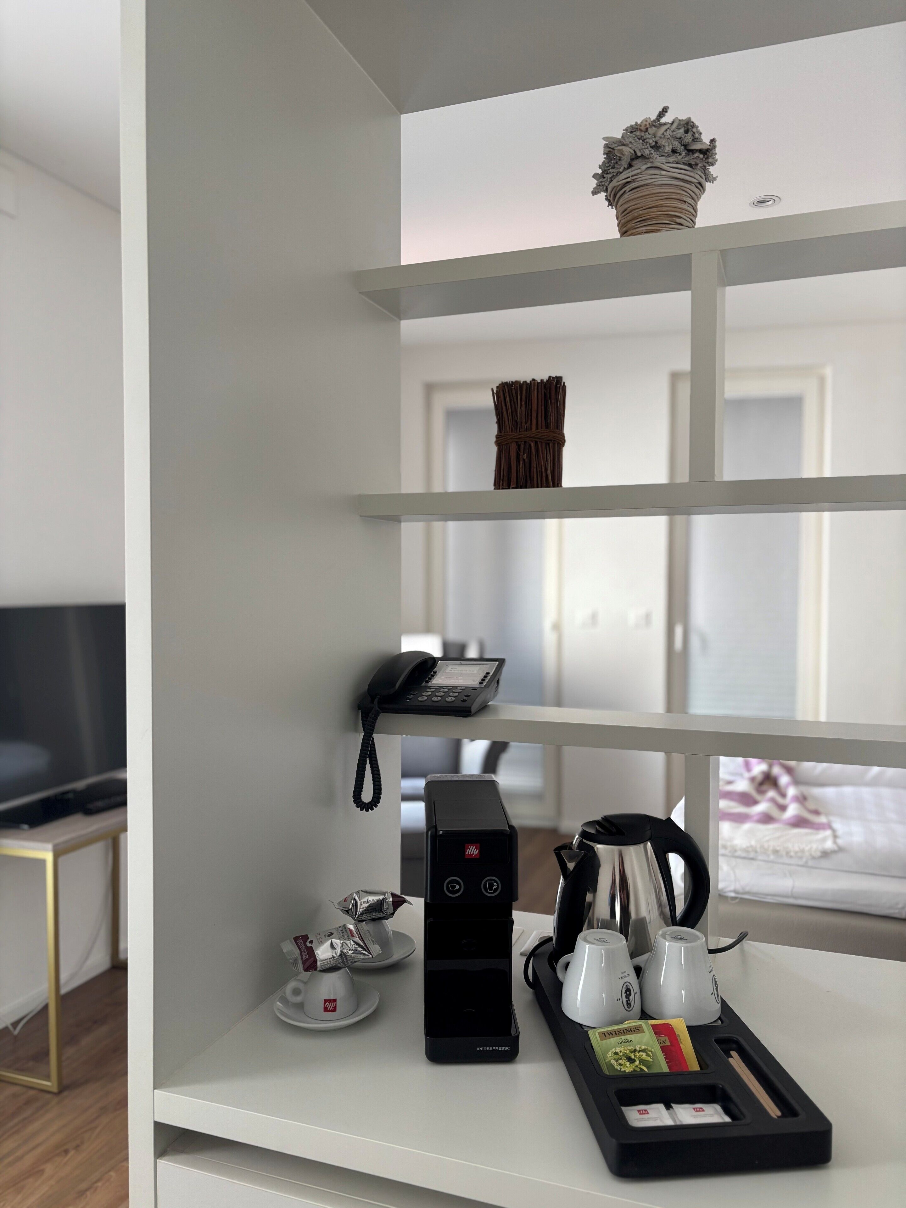 Habitación de hotel con una mesa blanca de consola, cafetera, teléfono y una cesta en los estantes.