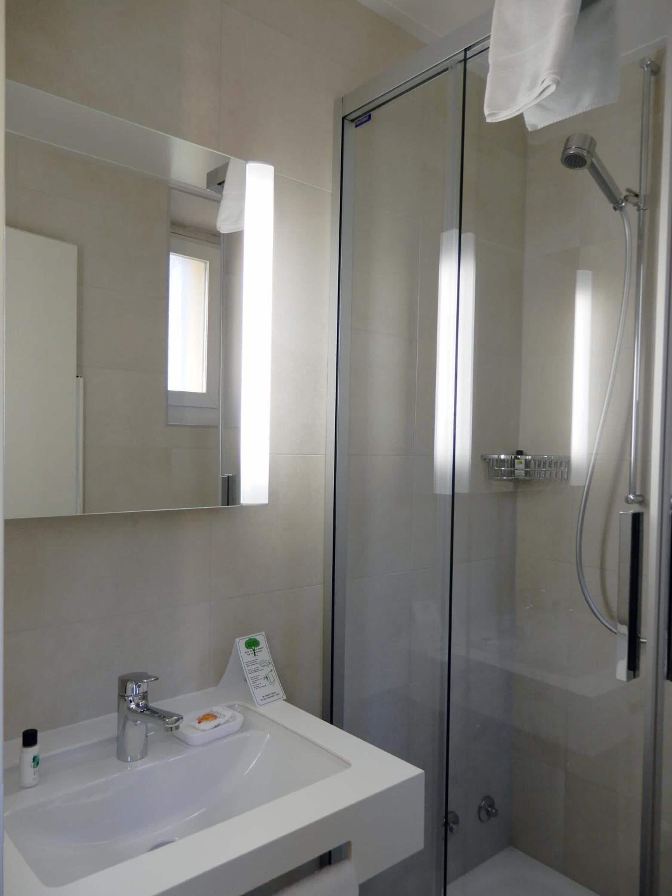 A modern bathroom with a glass shower enclosure, a white sink with a mirror above it, and a small window providing natural light.